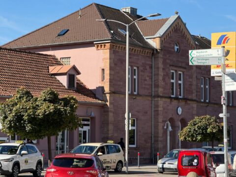 A pink brick building with tiled roof, street signs, and parked taxis along a sunny street.