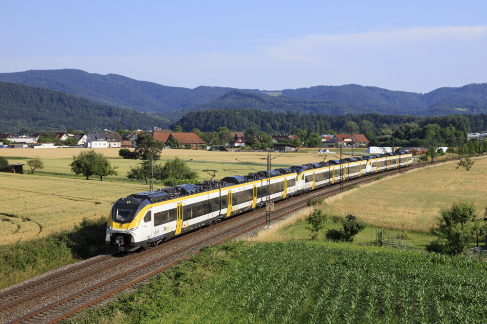 Passenger train with a yellow and white livery speeds along tracks through a rural countryside with houses, fields, and forested hills under a blue sky.