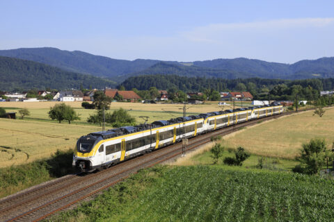 Passenger train with a yellow and white livery speeds along tracks through a rural countryside with houses, fields, and forested hills under a blue sky.