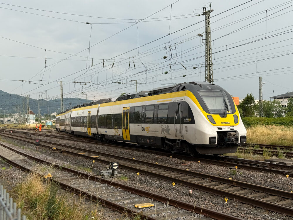 Modern yellow-and-gray passenger train on multiple tracks under extensive overhead power lines, with two workers in orange safety vests nearby.