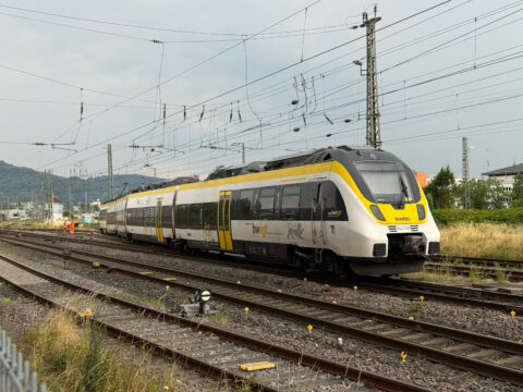 Modern yellow-and-gray passenger train on multiple tracks under extensive overhead power lines, with two workers in orange safety vests nearby.