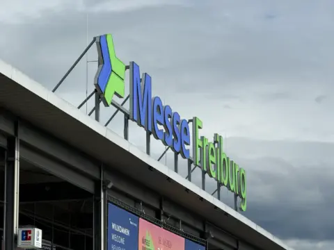 Close-up of blue and green 'Messe' sign on a modern convention center under a cloudy sky.