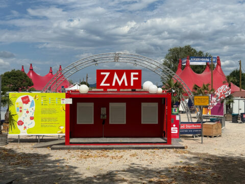 Festival booth with a red structure and a large white ZMF sign, red tents in the background.