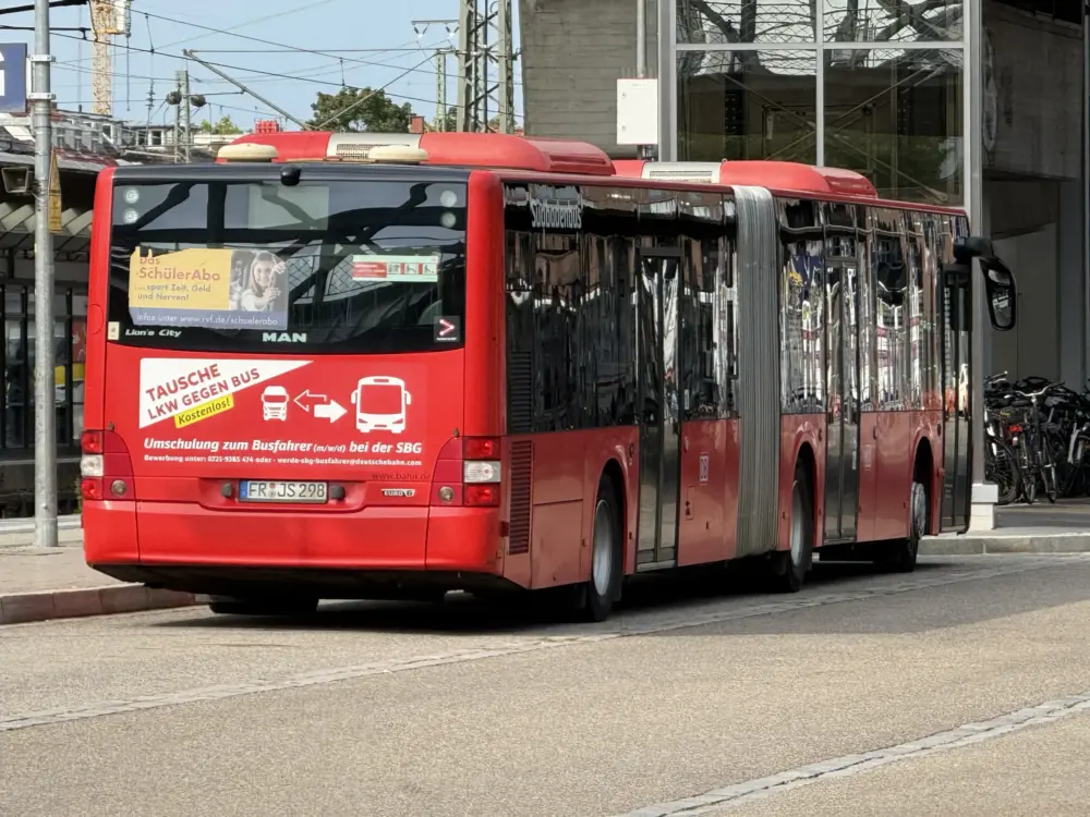 Red city bus stopped on a street, with posters on its back and a glossy, reflective side panel in an urban setting.