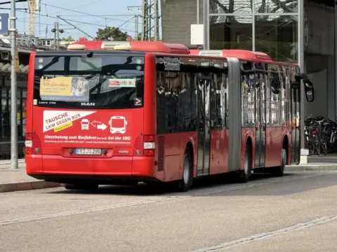 Red city bus stopped on a street, with posters on its back and a glossy, reflective side panel in an urban setting.